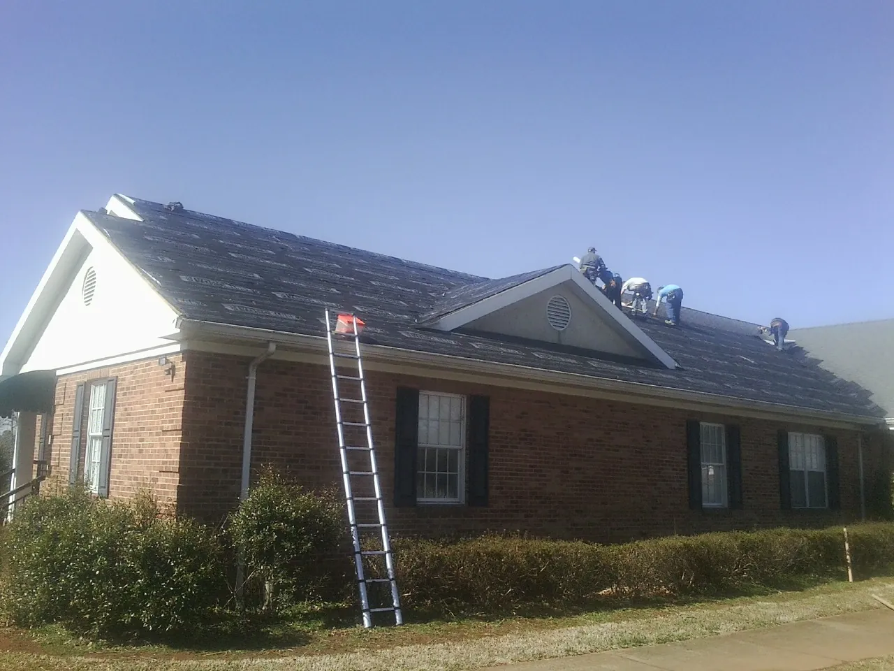 Crew tearing shingles off a brick ranch roof with a ladder set — Emergency Roof Repair work by our crew in Gilbert